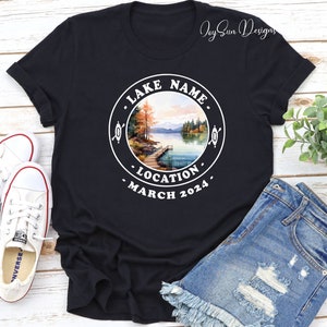 May include: Black t-shirt with a white circle graphic featuring a lake scene with trees, a dock, and the text "LAKE NAME", "LOCATION", and "MARCH 2024".