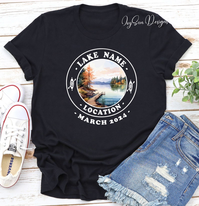 May include: Black t-shirt with a white circle graphic featuring a lake scene with trees, a dock, and the text "LAKE NAME", "LOCATION", and "MARCH 2024".