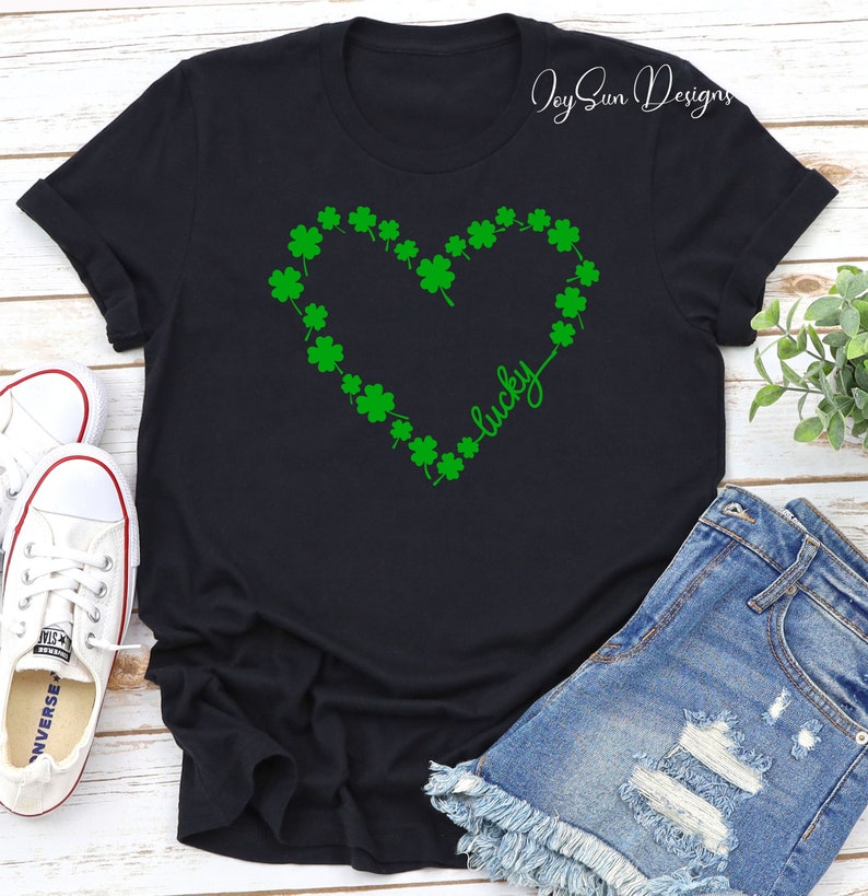 May include: Black t-shirt with a green shamrock heart design and the word "lucky" in cursive. The shirt is styled with denim shorts and white trainers. The image has a St. Patrick's Day theme.