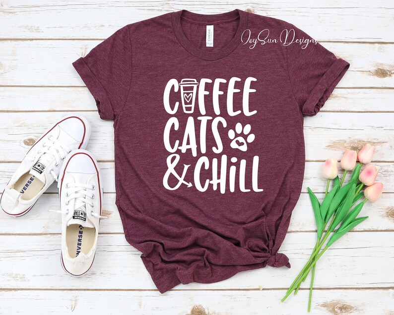 Puede incluir: Una camiseta de color granate con texto blanco que dice "Coffee Cats & Chill". El texto incluye un icono de taza de caf&eacute;, una huella de pata y un coraz&oacute;n. La camiseta est&aacute; colocada sobre una superficie de madera blanca con un par de zapatillas blancas y un ramo de tulipanes rosas.