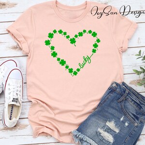 May include: A pale pink t-shirt with a green shamrock heart design and the word "lucky" in script. The shirt is styled with denim shorts and white trainers. The image has a St. Patrick's Day theme.