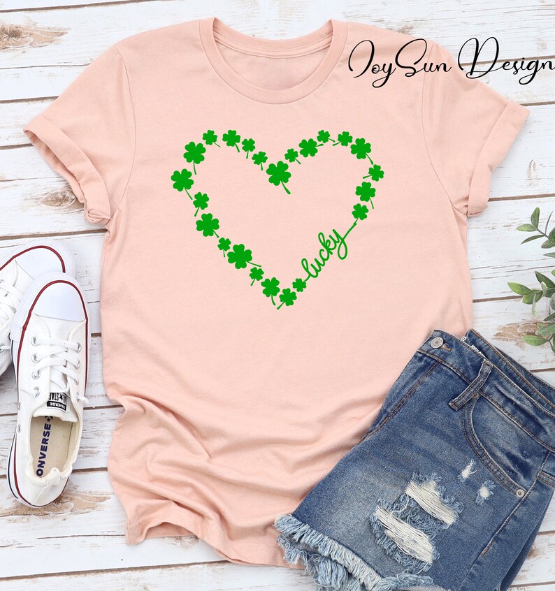 May include: A pale pink t-shirt with a green shamrock heart design and the word "lucky" in script. The shirt is styled with denim shorts and white trainers. The image has a St. Patrick's Day theme.