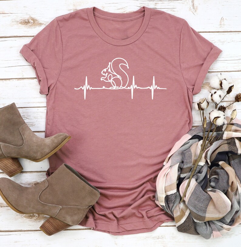 May include: Dusty rose-colored t-shirt featuring a white graphic of a squirrel within a heartbeat line. The shirt is styled with brown suede ankle boots and a patterned scarf. The design is simple and eye-catching.