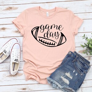 May include: A pink t-shirt with a black graphic of a football and the text "game day". The shirt is laying on a white wooden surface with a pair of white sneakers and blue denim shorts.