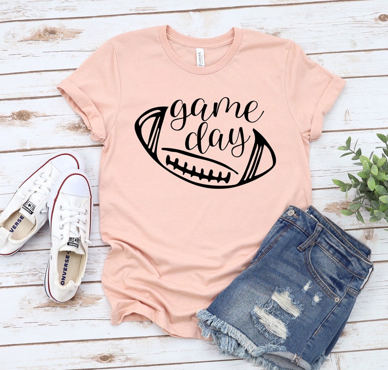 May include: A pink t-shirt with a black graphic of a football and the text "game day". The shirt is laying on a white wooden surface with a pair of white sneakers and blue denim shorts.