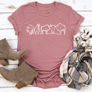 May include: Dusty rose-colored t-shirt featuring a white line drawing of several bison. The shirt is styled with brown suede ankle boots, a plaid scarf, and cotton stems. The shirt has a crew neck and short sleeves.