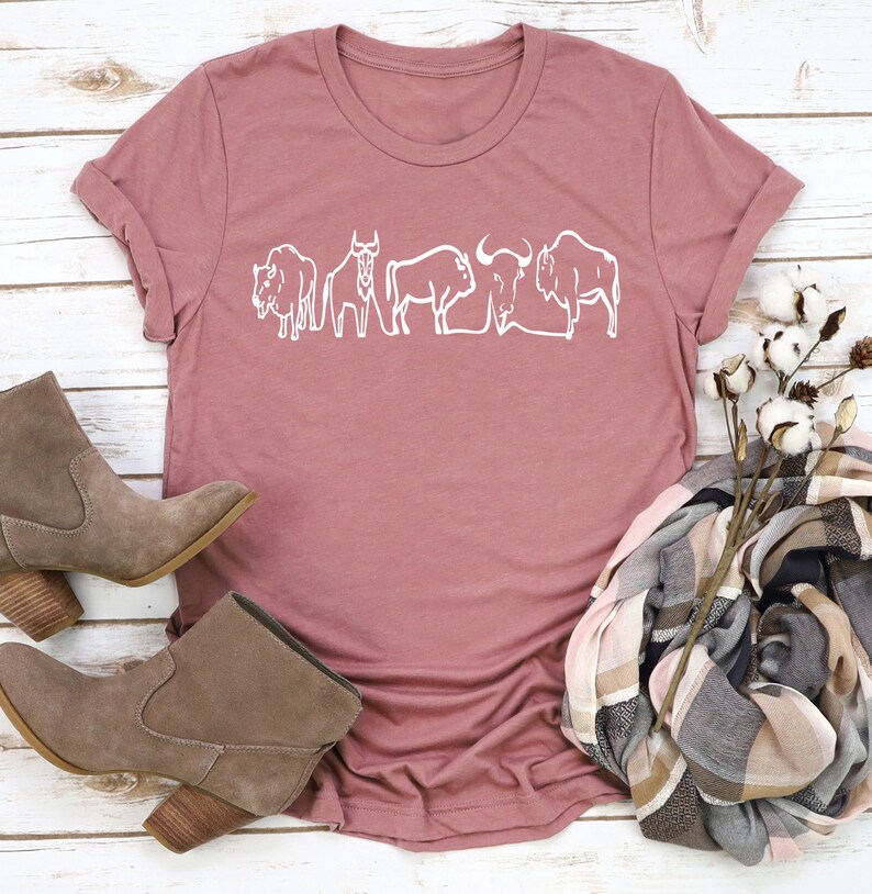 May include: Dusty rose-colored t-shirt featuring a white line drawing of several bison. The shirt is styled with brown suede ankle boots, a plaid scarf, and cotton stems. The shirt has a crew neck and short sleeves.