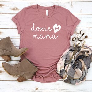 K&ouml;nnte beinhalten: Ein staubiges rosafarbenes T-Shirt mit dem Text "doxie mama" in wei&szlig;en Buchstaben. Eine wei&szlig;e Silhouette eines Dackels befindet sich in einem Herzen.
