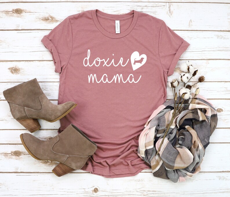 K&ouml;nnte beinhalten: Ein staubiges rosafarbenes T-Shirt mit dem Text "doxie mama" in wei&szlig;en Buchstaben. Eine wei&szlig;e Silhouette eines Dackels befindet sich in einem Herzen.