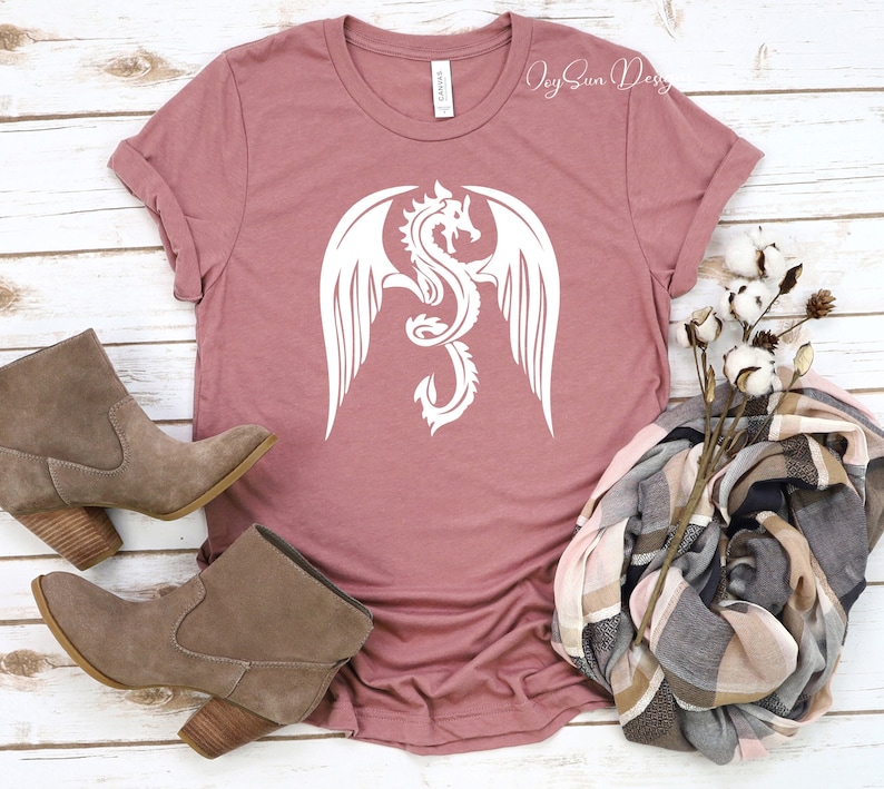 May include: A mauve t-shirt with a white graphic of a dragon with wings spread. The t-shirt is laying on a white wooden surface with a pair of brown suede ankle boots, a plaid scarf, and a cotton branch.