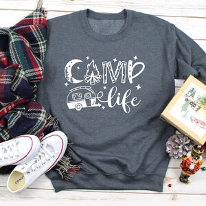 Pu&ograve; includere: Una felpa grigia scura con il testo "Camp life" in bianco. Il design include un fal&ograve;, una luna, stelle e un camper.