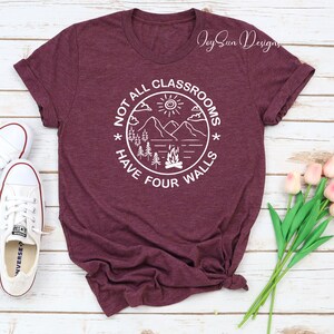 May include: A maroon t-shirt with a white graphic design that says "Not all classrooms have four walls". The graphic features a mountain scene with a campfire, trees, and a sun.