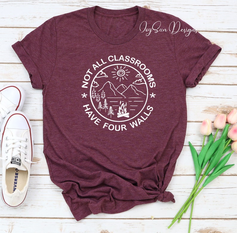 May include: A maroon t-shirt with a white graphic design that says "Not all classrooms have four walls". The graphic features a mountain scene with a campfire, trees, and a sun.