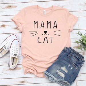 May include: A pink t-shirt with the text "MAMA CAT" in black lettering. The text is accompanied by a stylized cat face with whiskers and a heart-shaped nose.