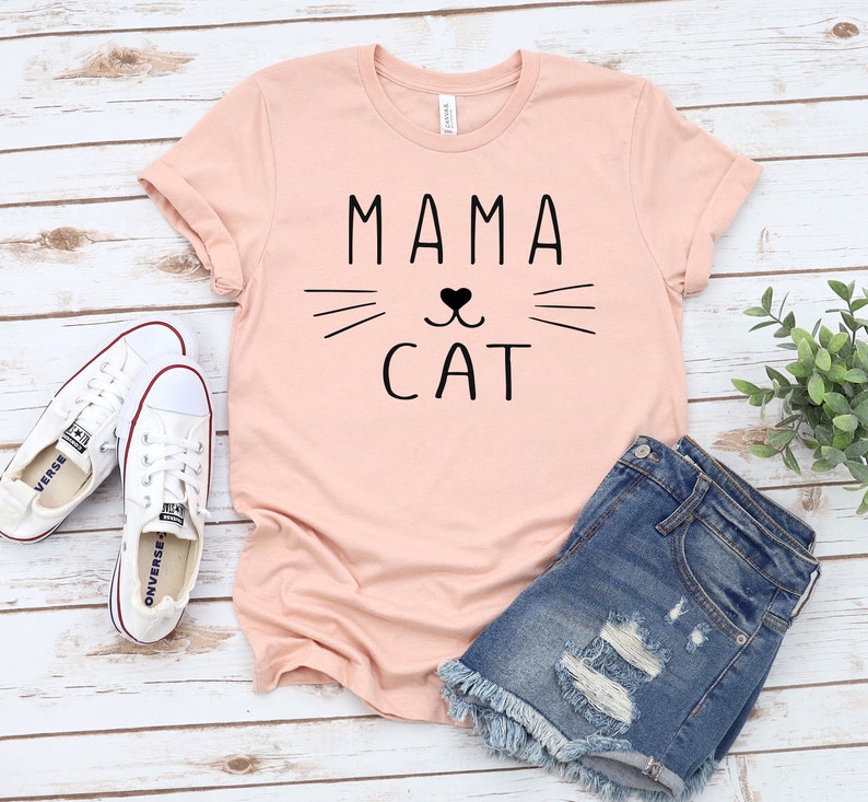 May include: A pink t-shirt with the text "MAMA CAT" in black lettering. The text is accompanied by a stylized cat face with whiskers and a heart-shaped nose.
