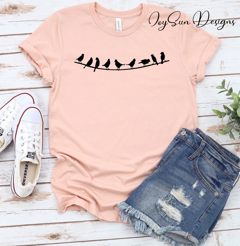 Peut inclure: Un t-shirt rose avec un motif de ligne d'oiseaux noirs sur un fil. Le texte "Joy Sun Designs" est dans le coin sup&eacute;rieur droit du t-shirt.