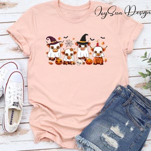 May include: A light pink t-shirt featuring a Halloween-themed graphic. The graphic depicts four Highland cows dressed in costumes, including witch hats and ghost costumes, with pumpkins, bats, and a spiderweb. The shirt is paired with denim shorts and white Converse sneakers.