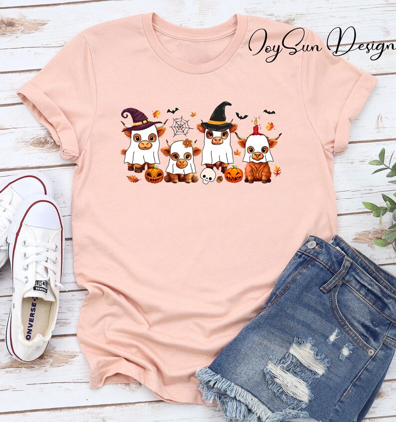May include: A light pink t-shirt featuring a Halloween-themed graphic. The graphic depicts four Highland cows dressed in costumes, including witch hats and ghost costumes, with pumpkins, bats, and a spiderweb. The shirt is paired with denim shorts and white Converse sneakers.