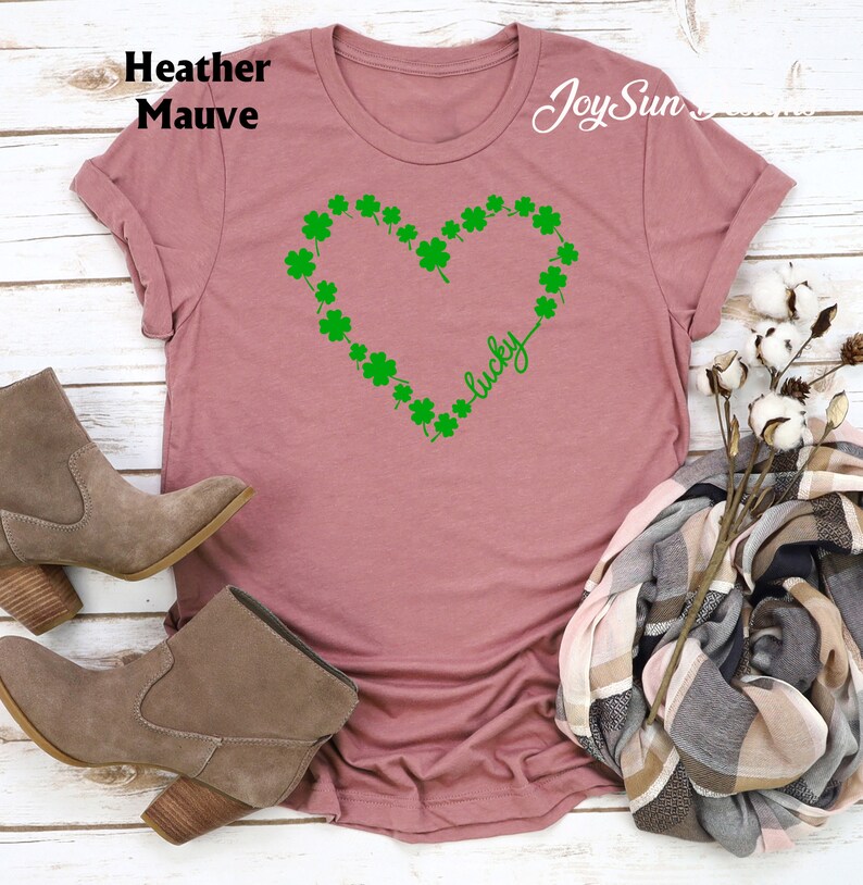 May include: Mauve t-shirt with a green shamrock heart design and the word "lucky". The shirt is a soft, casual style. The image also shows a pair of brown suede boots and a scarf.