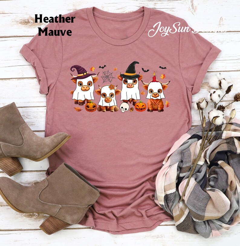 May include: Heather Mauve t-shirt featuring a Halloween-themed graphic. The design includes four cartoon cows dressed in costumes, pumpkins, a spider web, and the text "Joy Sun".