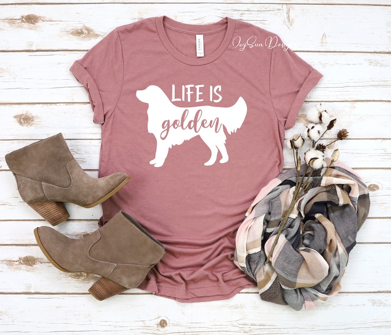 May include: A dusty rose t-shirt with a white silhouette of a golden retriever and the text "Life is golden". The shirt is laying on a white wooden surface with two brown suede ankle boots and a plaid scarf.