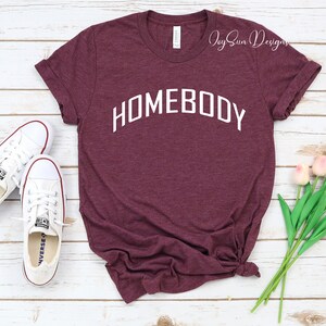 K&ouml;nnte beinhalten: Burgunderfarbenes T-Shirt mit dem Wort "HOMEBODY" in Wei&szlig;, das sich bogenf&ouml;rmig &uuml;ber die Brust spannt. Wei&szlig;e Turnschuhe befinden sich links vom Shirt. Ein kleiner Strau&szlig; rosa Tulpen befindet sich rechts. Das Shirt hat hochgekrempelte &Auml;rmel.