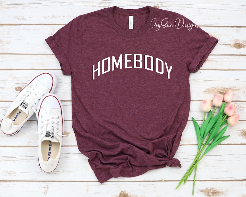 K&ouml;nnte beinhalten: Burgunderfarbenes T-Shirt mit dem Wort "HOMEBODY" in Wei&szlig;, das sich bogenf&ouml;rmig &uuml;ber die Brust spannt. Wei&szlig;e Turnschuhe befinden sich links vom Shirt. Ein kleiner Strau&szlig; rosa Tulpen befindet sich rechts. Das Shirt hat hochgekrempelte &Auml;rmel.