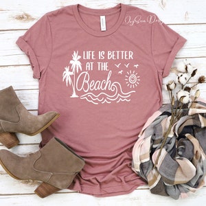 May include: A mauve t-shirt with the text "Life is better at the beach" in white. The design includes palm trees, a sun, waves, and birds. The t-shirt is laid flat on a white wooden surface with a pair of brown ankle boots and a plaid scarf.