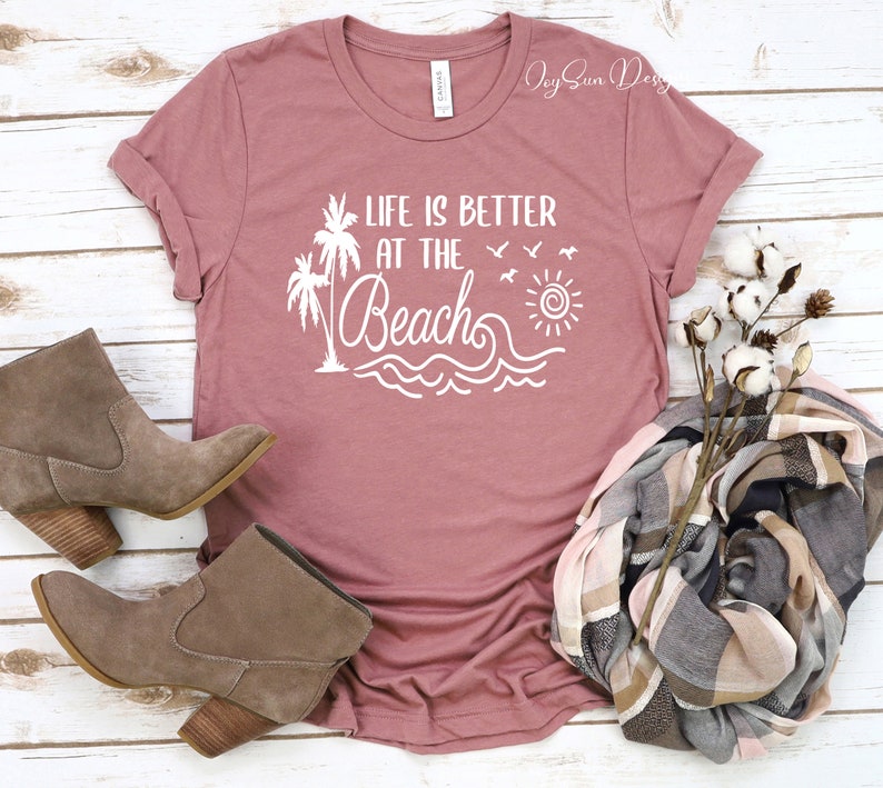May include: A mauve t-shirt with the text "Life is better at the beach" in white. The design includes palm trees, a sun, waves, and birds. The t-shirt is laid flat on a white wooden surface with a pair of brown ankle boots and a plaid scarf.