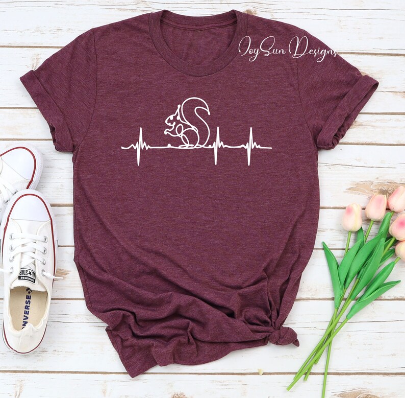 May include: Maroon t-shirt featuring a white graphic of a squirrel within a heartbeat line design. The text "Joy Sun Designs" is printed in cursive on the upper right. White sneakers and pink tulips are in the background.