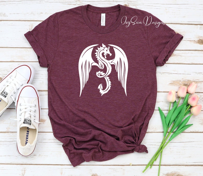 May include: A maroon t-shirt with a white graphic of a dragon with wings spread. The text "Joy Sun Designs" is visible in the top right corner of the image.