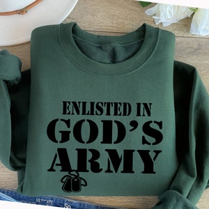 May include: A dark green sweatshirt with the text "Enlisted in God's Army" in black lettering. The text is in a bold, blocky font. There are two black dog tags hanging below the text.