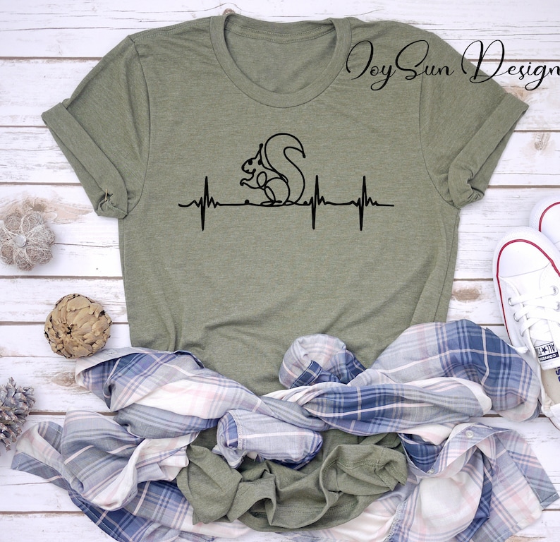 May include: Olive green t-shirt with a black line drawing of a squirrel and a heartbeat graphic. The shirt has rolled-up sleeves and is displayed with a plaid shirt and white sneakers.