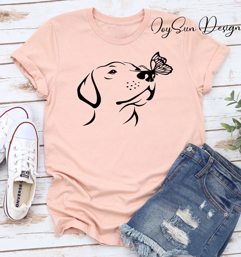 May include: A pink t-shirt with a black silhouette of a dog sniffing a butterfly. The text "Joy Sun Design" is in the top right corner of the shirt.