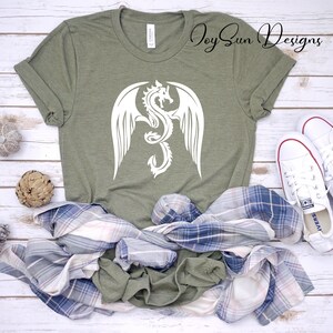 May include: A green t-shirt with a white dragon design. The dragon has wings and is curled up in a circle. The shirt is on a white wooden surface with a plaid shirt and white sneakers.