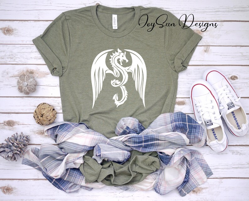 May include: A green t-shirt with a white dragon design. The dragon has wings and is curled up in a circle. The shirt is on a white wooden surface with a plaid shirt and white sneakers.