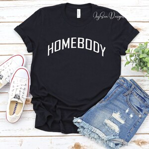 K&ouml;nnte beinhalten: Schwarzes T-Shirt mit dem wei&szlig;en Schriftzug "HOMEBODY" in Bogenform. Dazu Jeansshorts und wei&szlig;e Turnschuhe. Das Bild zeigt einen l&auml;ssigen, alltagstauglichen Kleidungsstil.
