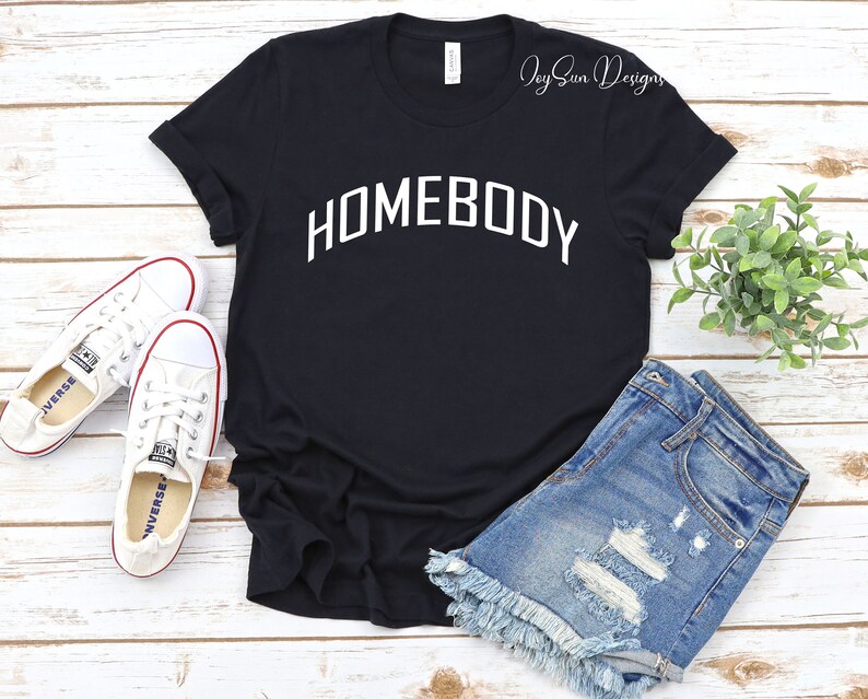 K&ouml;nnte beinhalten: Schwarzes T-Shirt mit dem wei&szlig;en Schriftzug "HOMEBODY" in Bogenform. Dazu Jeansshorts und wei&szlig;e Turnschuhe. Das Bild zeigt einen l&auml;ssigen, alltagstauglichen Kleidungsstil.