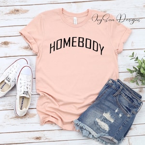 K&ouml;nnte beinhalten: Hellrosa T-Shirt mit dem schwarzen Wort "HOMEBODY" in Bogenform. Das Shirt wird mit Jeansshorts und wei&szlig;en Leinenschuhen kombiniert. Das Shirt ist von Joy Sun Designs.