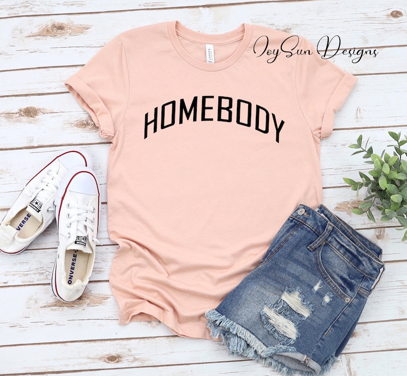 K&ouml;nnte beinhalten: Hellrosa T-Shirt mit dem schwarzen Wort "HOMEBODY" in Bogenform. Das Shirt wird mit Jeansshorts und wei&szlig;en Leinenschuhen kombiniert. Das Shirt ist von Joy Sun Designs.