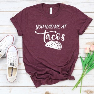 以下が含まれることがあります： 白地に「You had me at tacos」と書いてある、ワインレッドのTシャツ。白いアウトラインのタコスが描かれている。