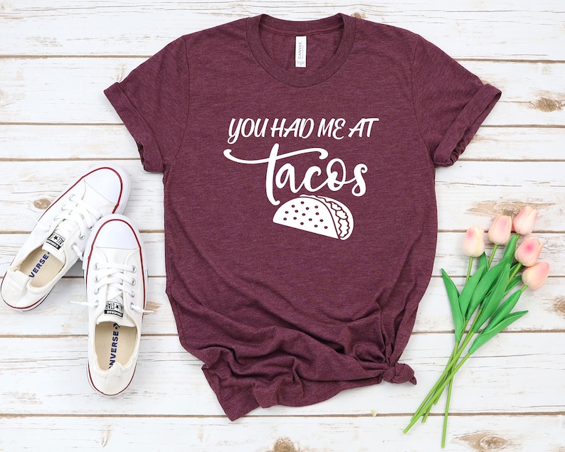 以下が含まれることがあります： 白地に「You had me at tacos」と書いてある、ワインレッドのTシャツ。白いアウトラインのタコスが描かれている。