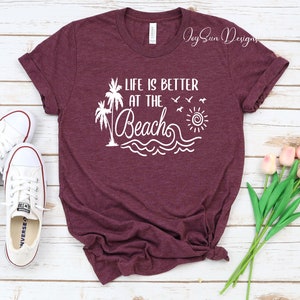 May include: A maroon t-shirt with the text "Life is better at the beach" in white. The design includes palm trees, a sun, and waves.