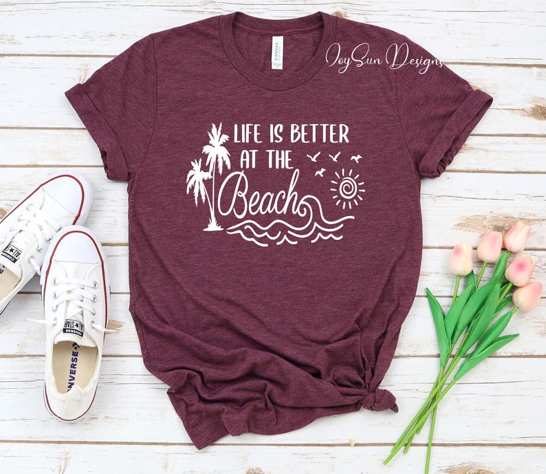 May include: A maroon t-shirt with the text "Life is better at the beach" in white. The design includes palm trees, a sun, and waves.