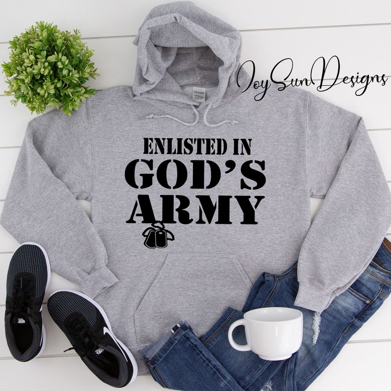 May include: A gray hooded sweatshirt with the text "Enlisted in God's Army" in black lettering. The sweatshirt has a pocket on the front and is shown on a white wooden surface with a pair of black sneakers, a white mug, and blue jeans.