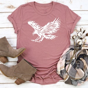 May include: A mauve t-shirt with a white graphic of a stylized eagle with flames for wings.