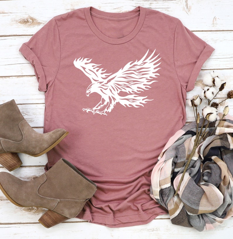 May include: A mauve t-shirt with a white graphic of a stylized eagle with flames for wings.
