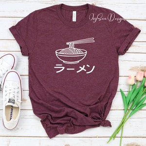 Puede incluir: Una camiseta de color granate con un contorno blanco de un cuenco de fideos ramen y palillos. La palabra japonesa para ramen, "ラーメン", est&aacute; escrita debajo del cuenco.