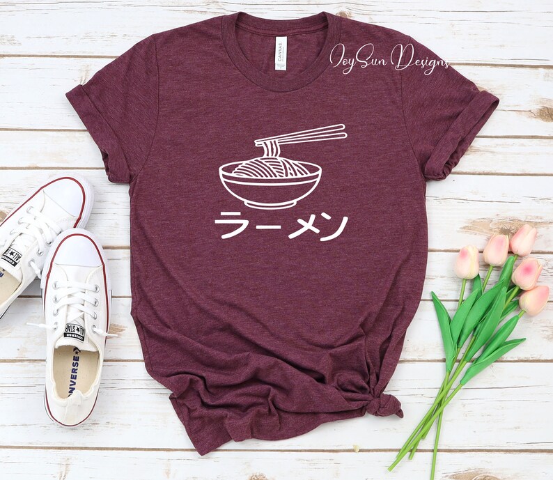 Puede incluir: Una camiseta de color granate con un contorno blanco de un cuenco de fideos ramen y palillos. La palabra japonesa para ramen, "ラーメン", est&aacute; escrita debajo del cuenco.