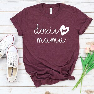 K&ouml;nnte beinhalten: Burgunderfarbenes T-Shirt mit wei&szlig;em Text "doxie mama" und einer wei&szlig;en Silhouette eines Dackels in einem Herzen.